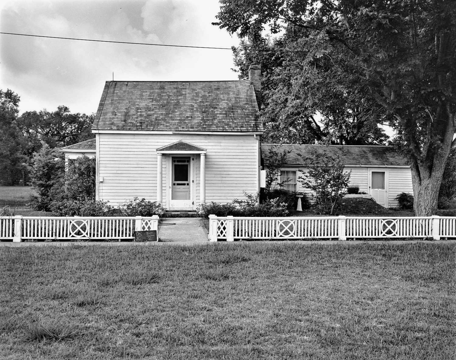 House with white siding and gray shingled roof, wooden fence, trees, and grass