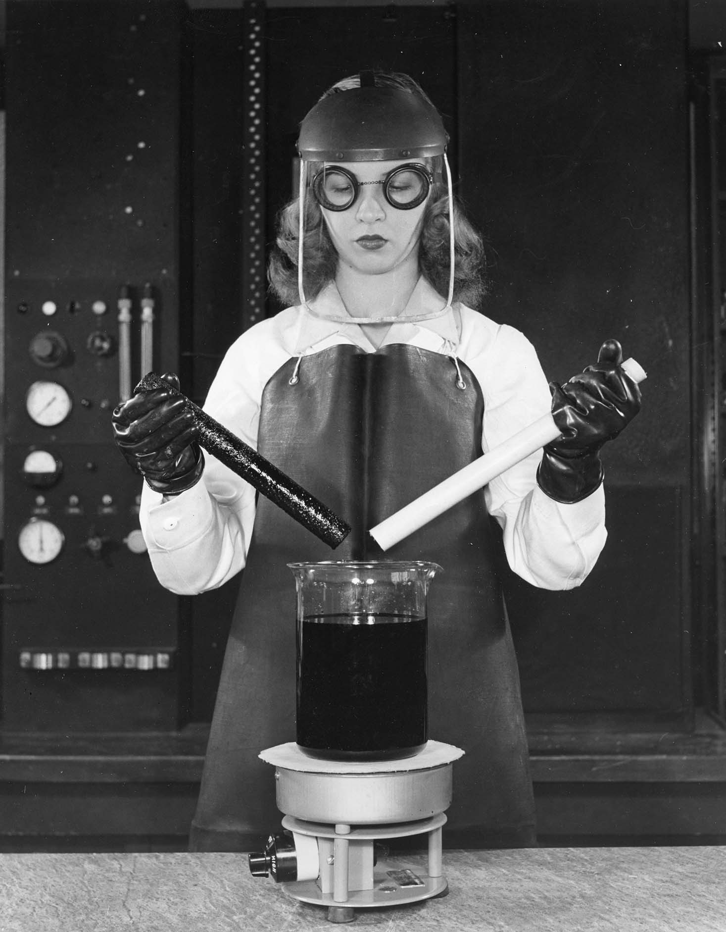 Woman in goggles, smock, and face shield in a lab holding plastic rods over a glass beaker of chemicals