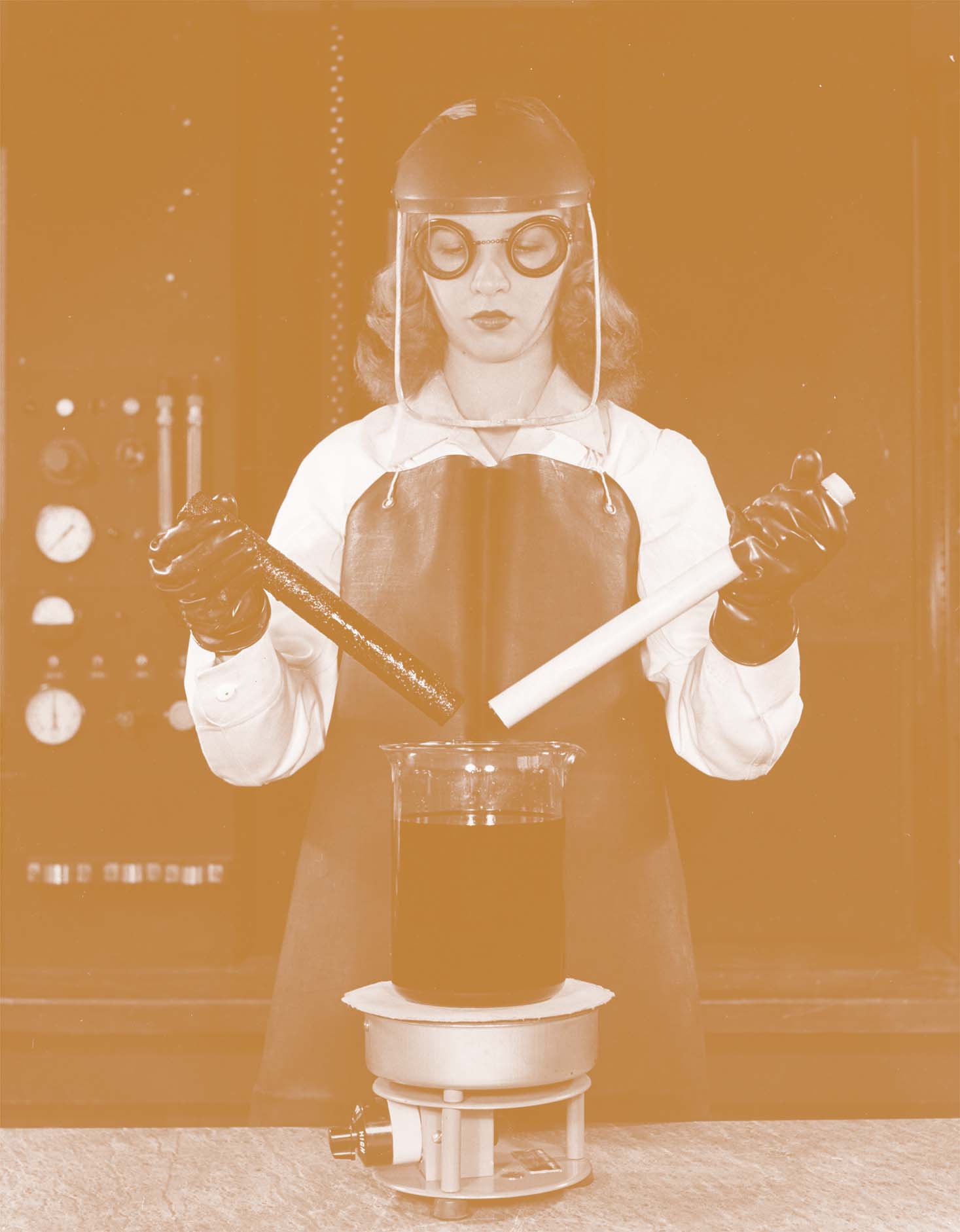 Woman in goggles, smock, and face shield in a lab holding plastic rods over a glass beaker of chemicals