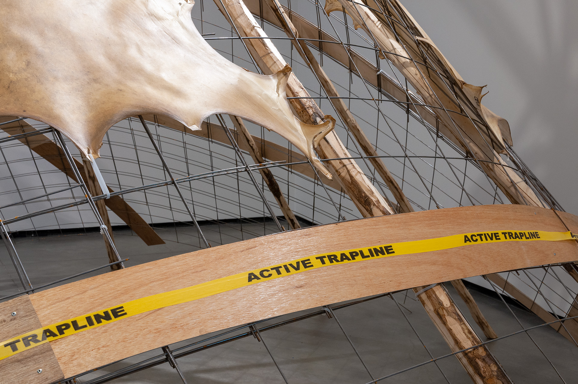 Detail of a wire mesh structure with animal hide and wood with the text “Active Trapline” across it