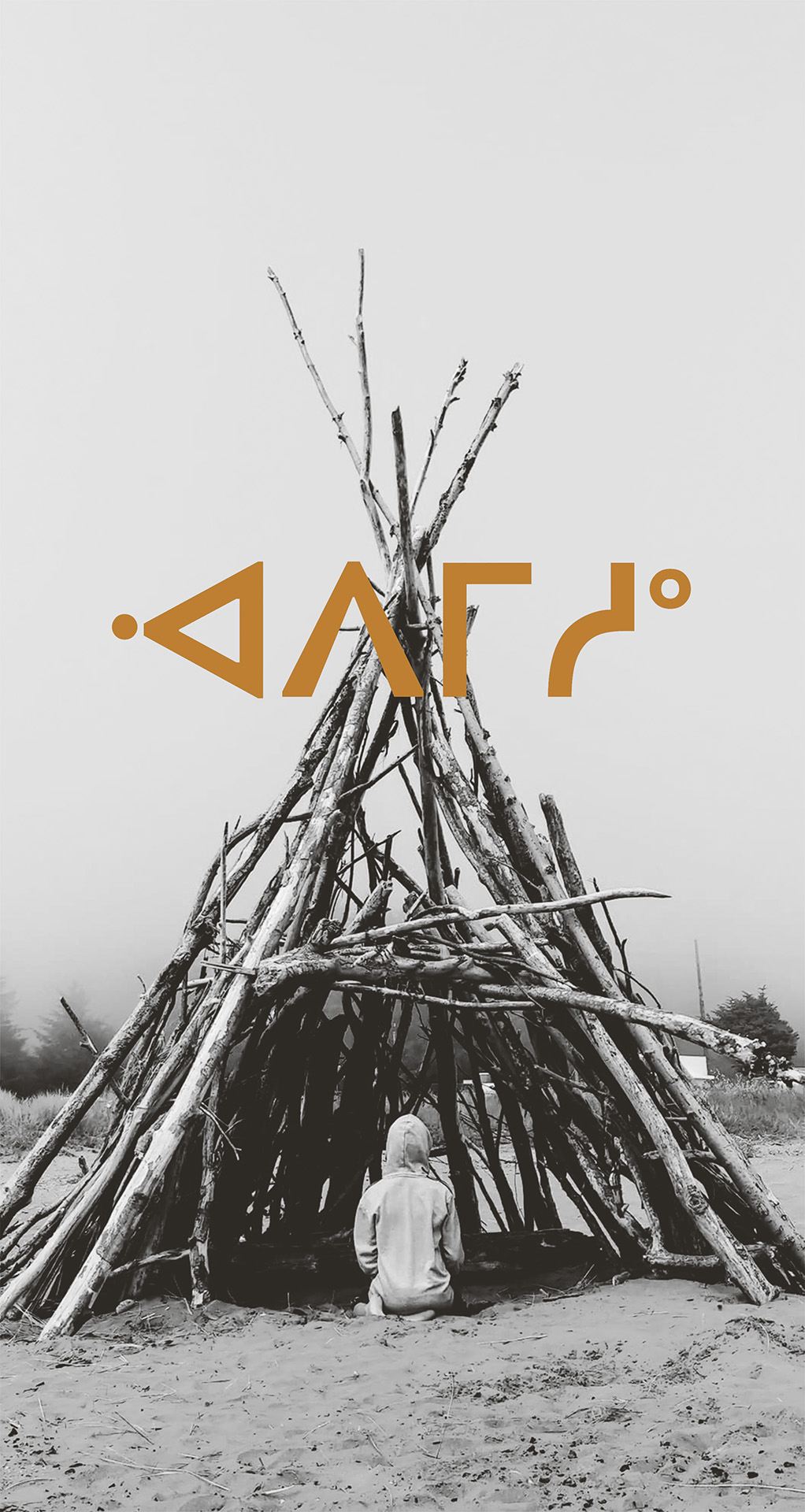 A person with their back to the camera sits in a wooden tipi on sand. Cree syllabics are overlayed